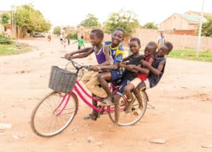 Hopeful, dignified children, in the context of their homes and neighborhoods. A group of boys, friends, buddies, children, youth, five boys pile all on top of a bicycle, bike, transportation, on a dirt road in their neighborhood street. The bike has a gray woven basket on it and the boys are smiling together.