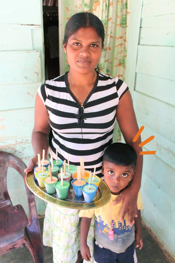Compassion's Sensitive Messaging Standards apply to this asset for external usage. See Compassion's Communications Manual (p. 22-26) https://maxplus.compassion.com/viewpicture.tlx?pictureid=22119320320 for the standards. 

Vanantha Rani, a CSP mother, adult woman, stands outside her home holding her young child, son, Imesh with one hand at his shoulder and carrying a tray with popsicles, ice pops, snacks, in the other hand.